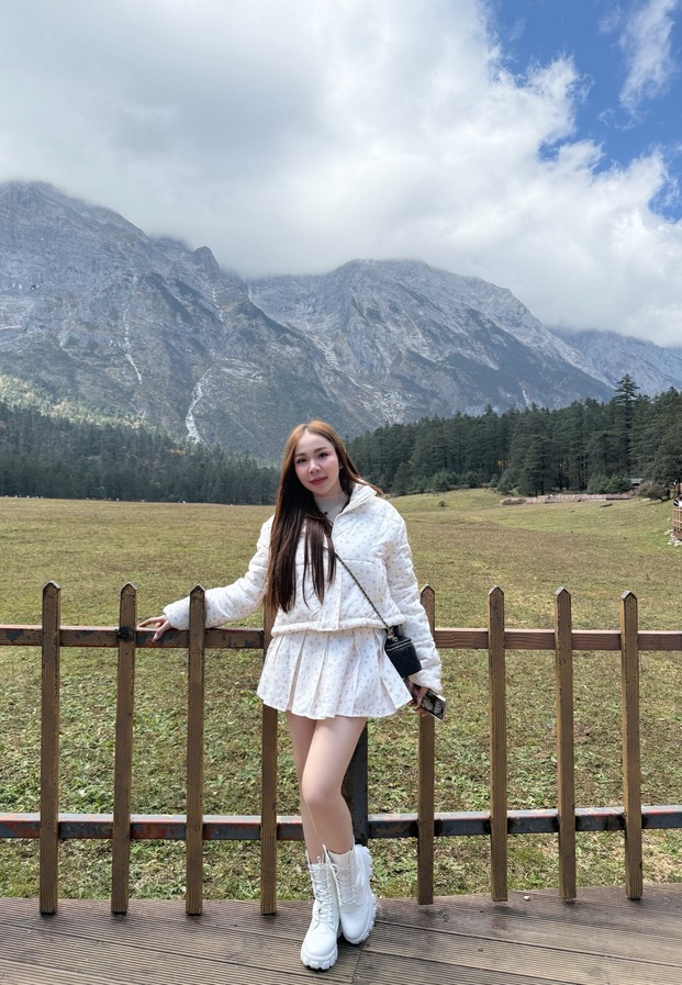 แจ็กเก็ตสีขาวพิงรั้วไม้หน้าภูเขาที่ลี่เจียง